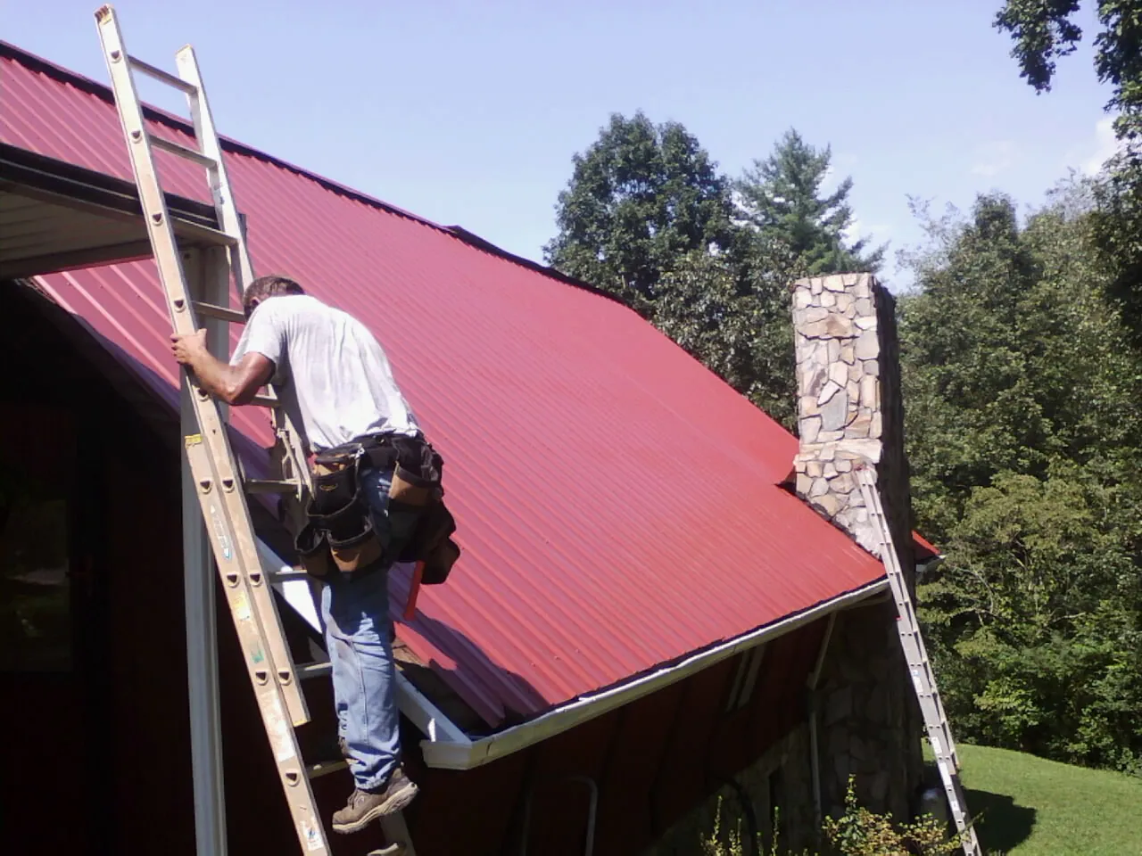 Roofer on a ladder beside a red metal roof and stone chimney — Soffit & Fascia Repair by Roofing Brothers in Bradley Beach
